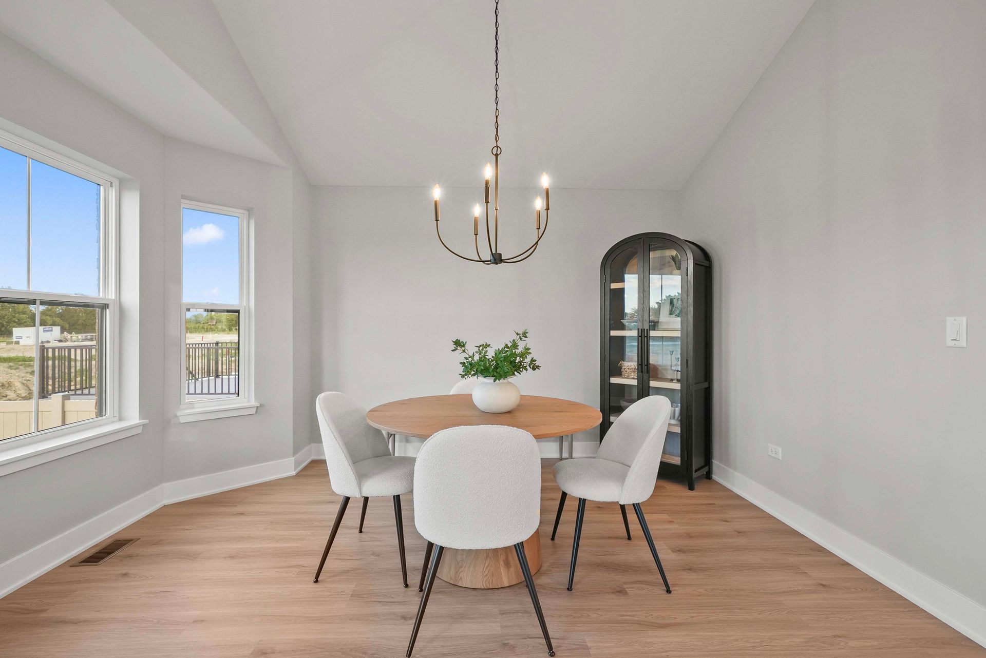 Bright dining room with round table, white chairs, chandelier, and black glass cabinet; large windows on left.