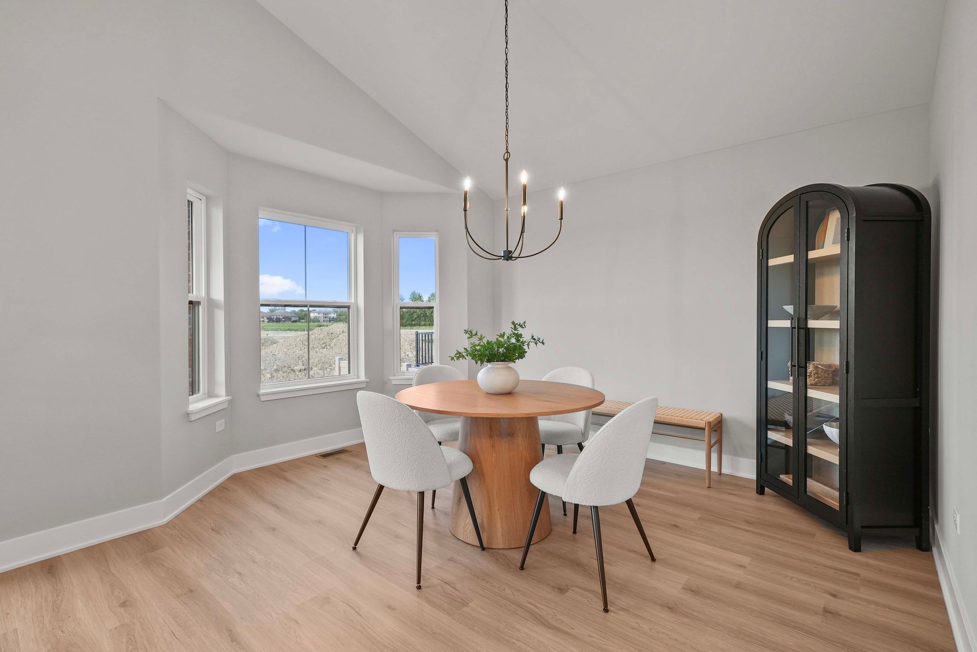 Bright dining room with round wooden table, white chairs, black cabinet, and large windows.