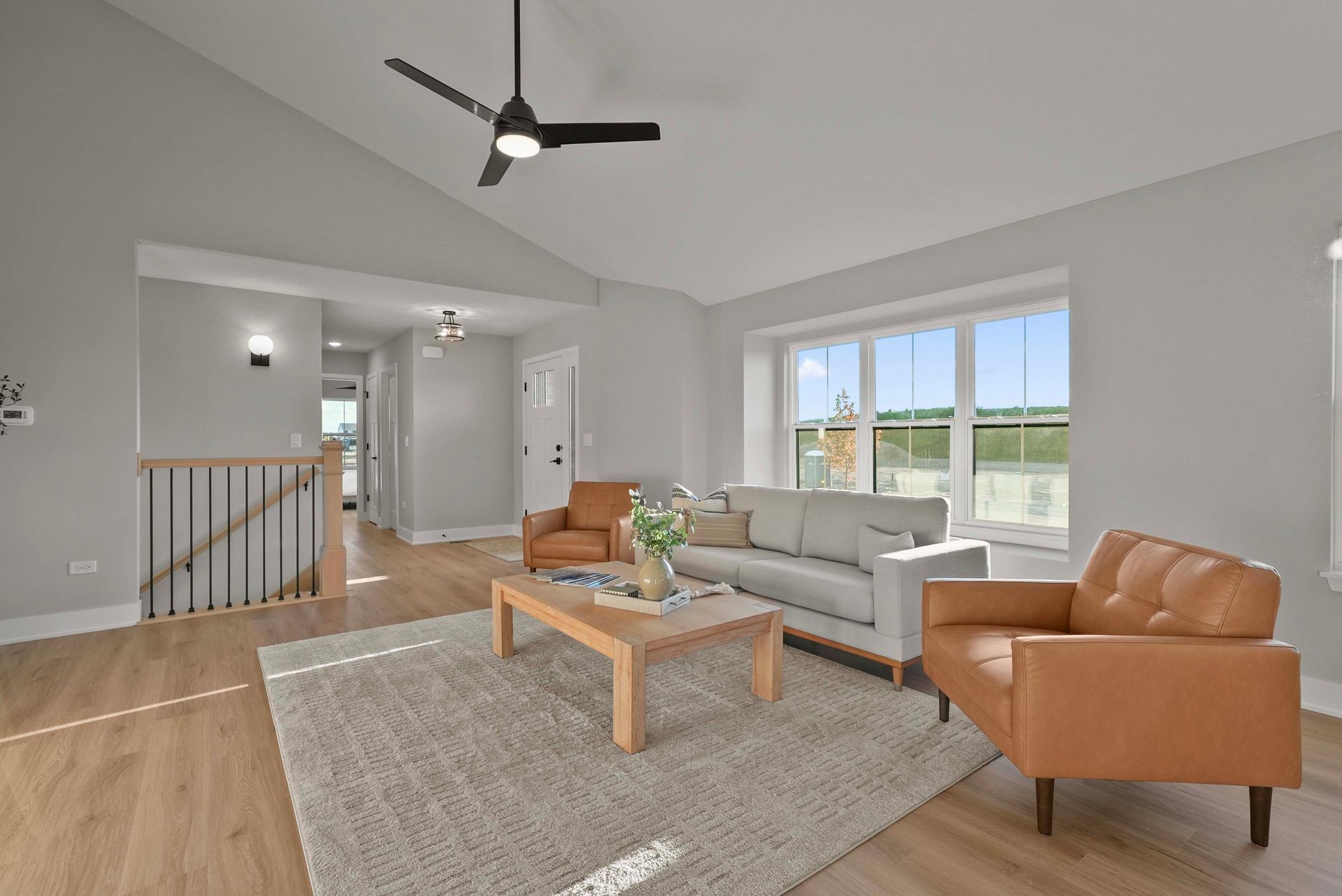 Bright living room with gray sofa, tan chairs, wood coffee table, and large windows under a ceiling fan