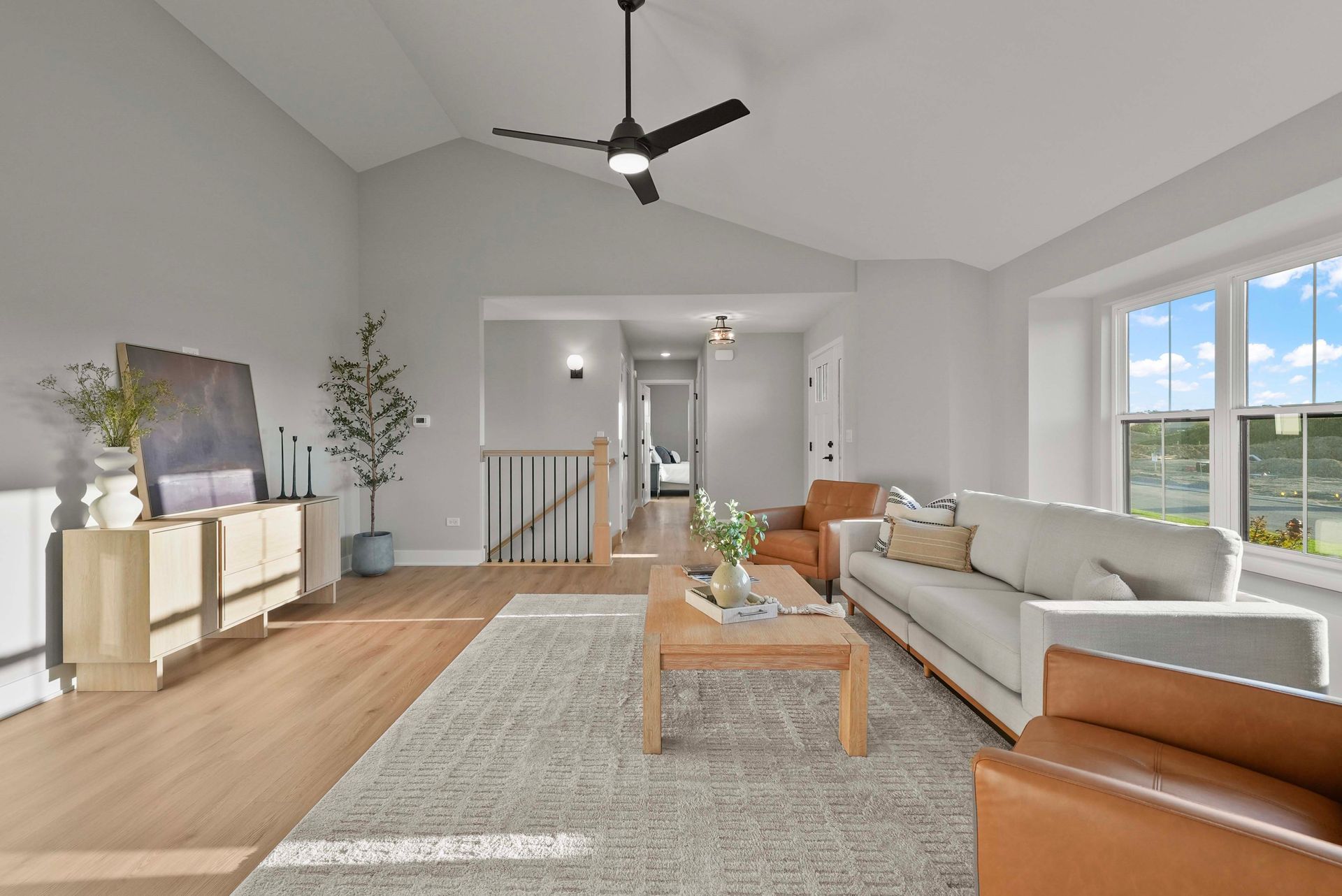 Bright modern living room with gray sofa, tan chairs, rug, wood floor, and large windows