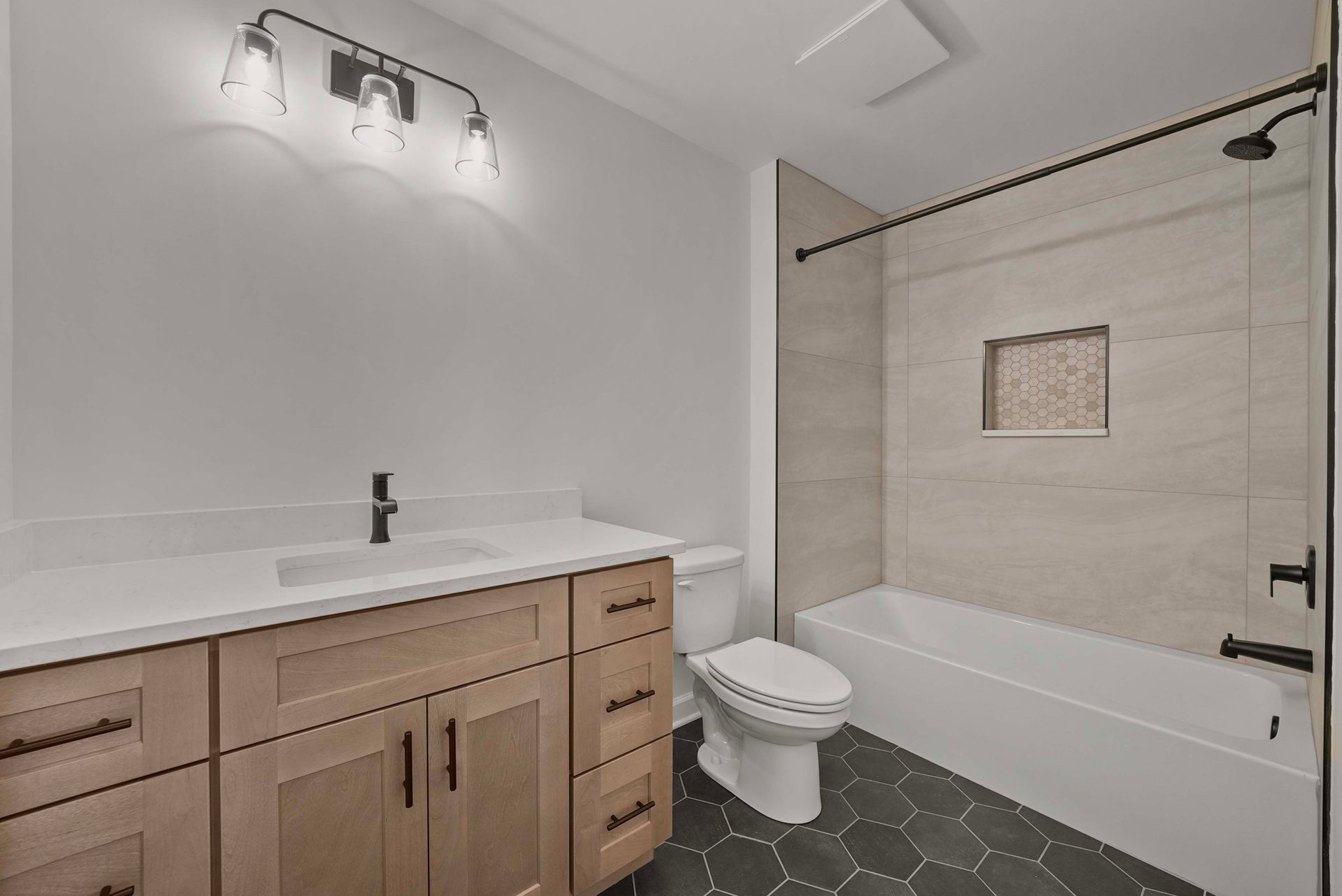 Modern bathroom with white tub and shower, wood vanity, dark tile floor, and wall light.