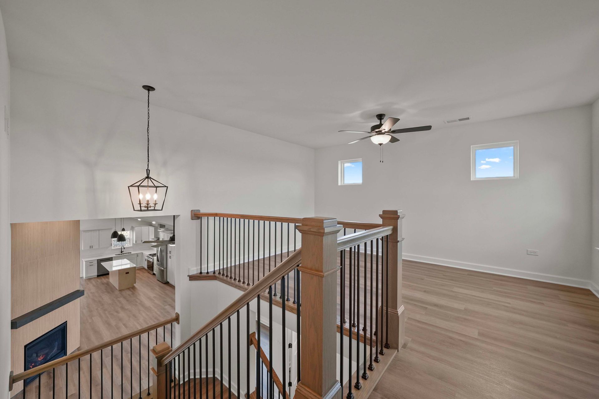 Bright open loft with white walls, ceiling fan, hardwood floors, railing, and a view to the kitchen below.