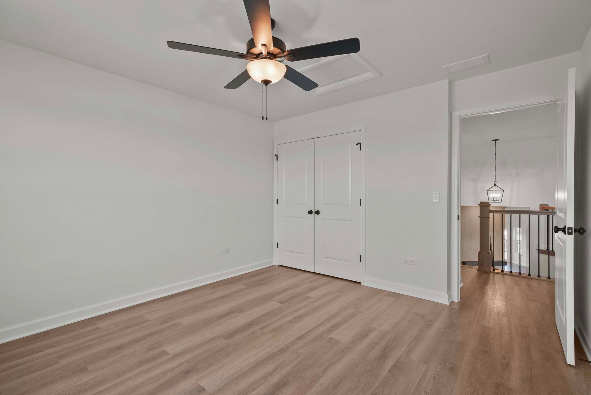 Empty bedroom with light wood floors, white walls, ceiling fan, and mirrored closet doors.