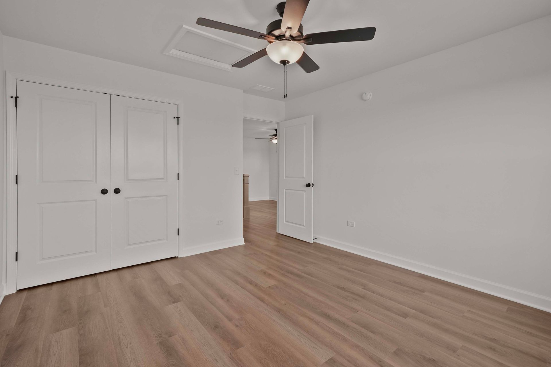 Empty white bedroom with wood flooring, ceiling fan, and double closet doors