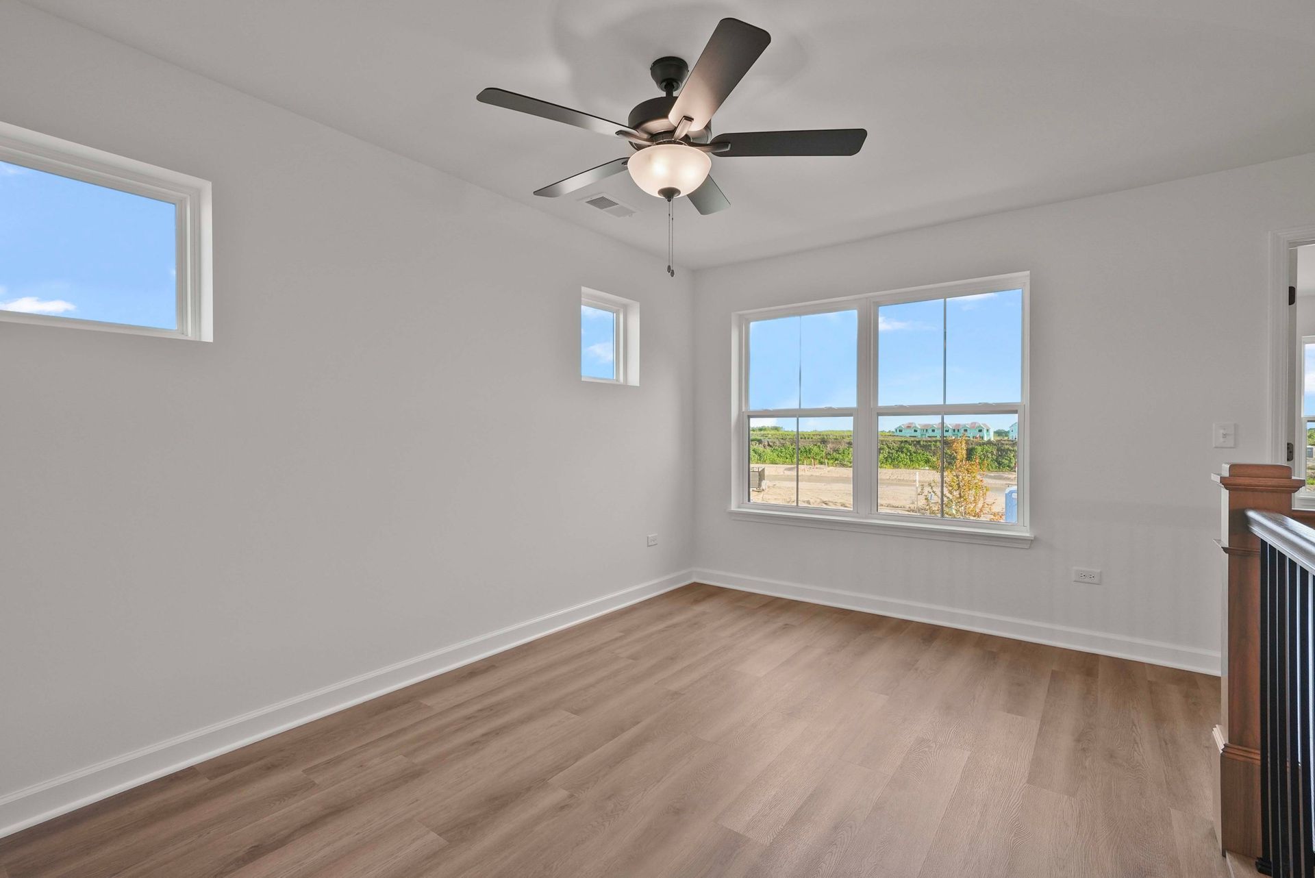 Empty white bedroom with wood floors, ceiling fan, and two windows overlooking a field.