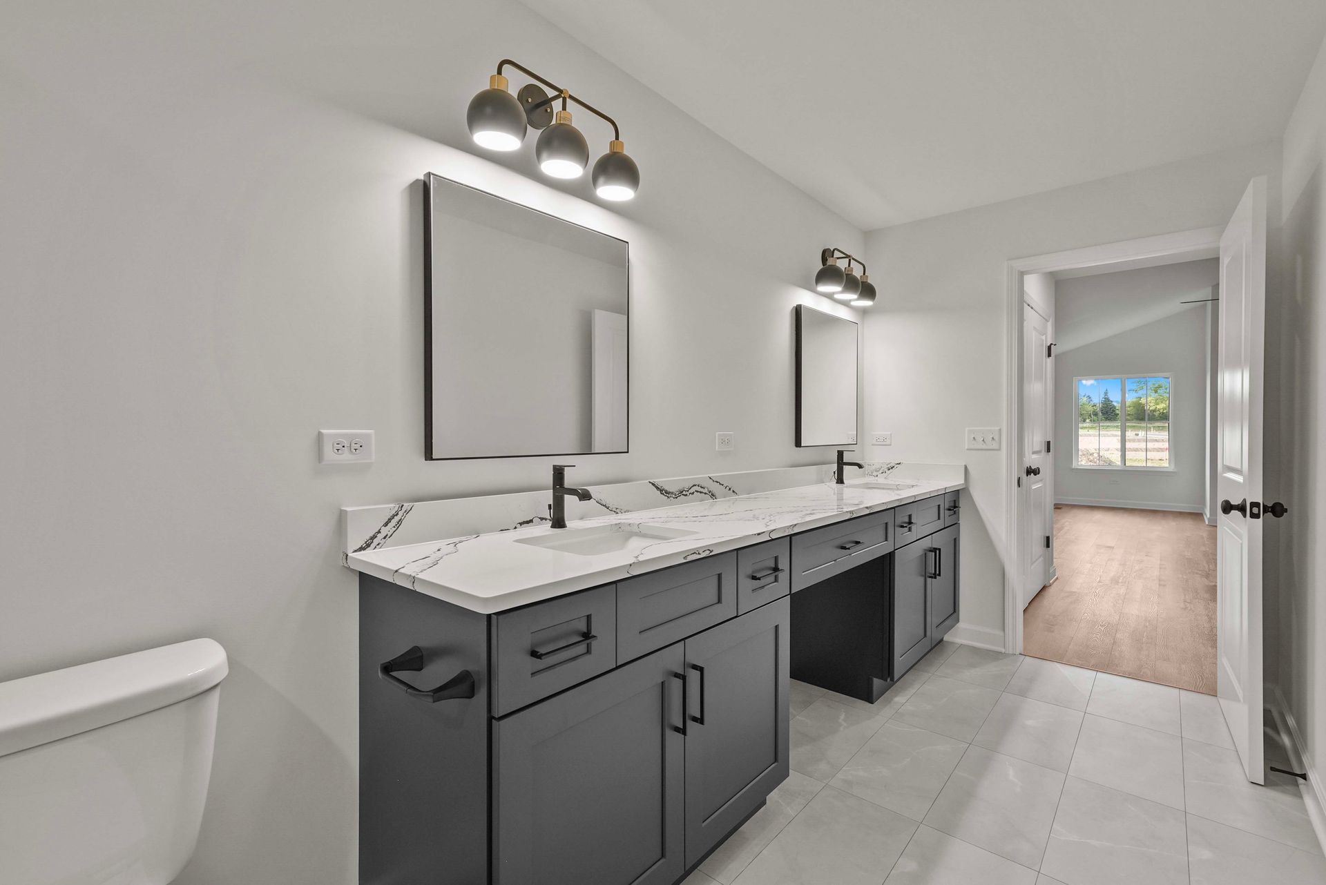 Modern bathroom vanity with double sinks, gray cabinets, and mirrors under wall lights, leading to a bedroom.