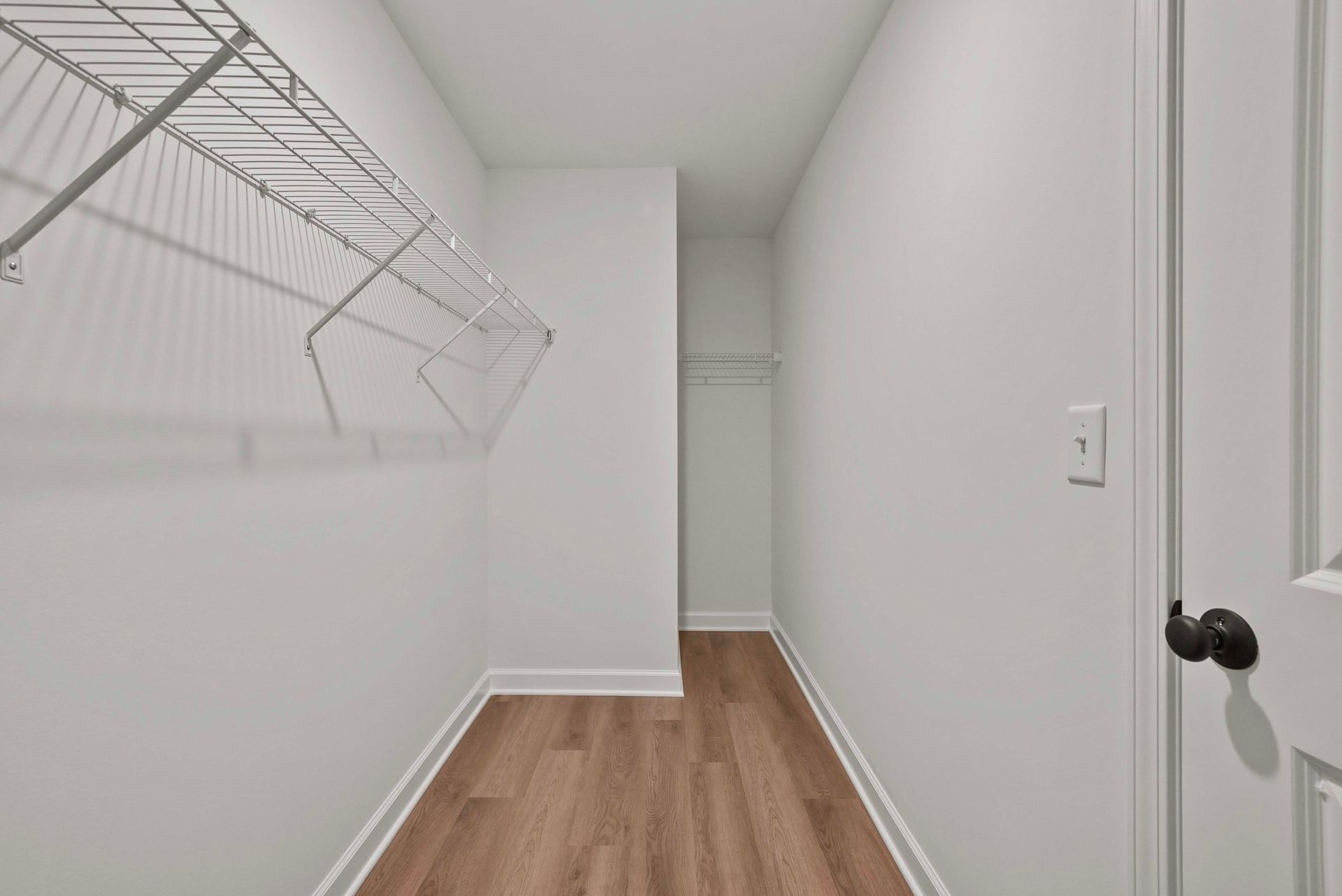 Empty white closet with wire shelving and wood flooring, viewed down a narrow hallway.