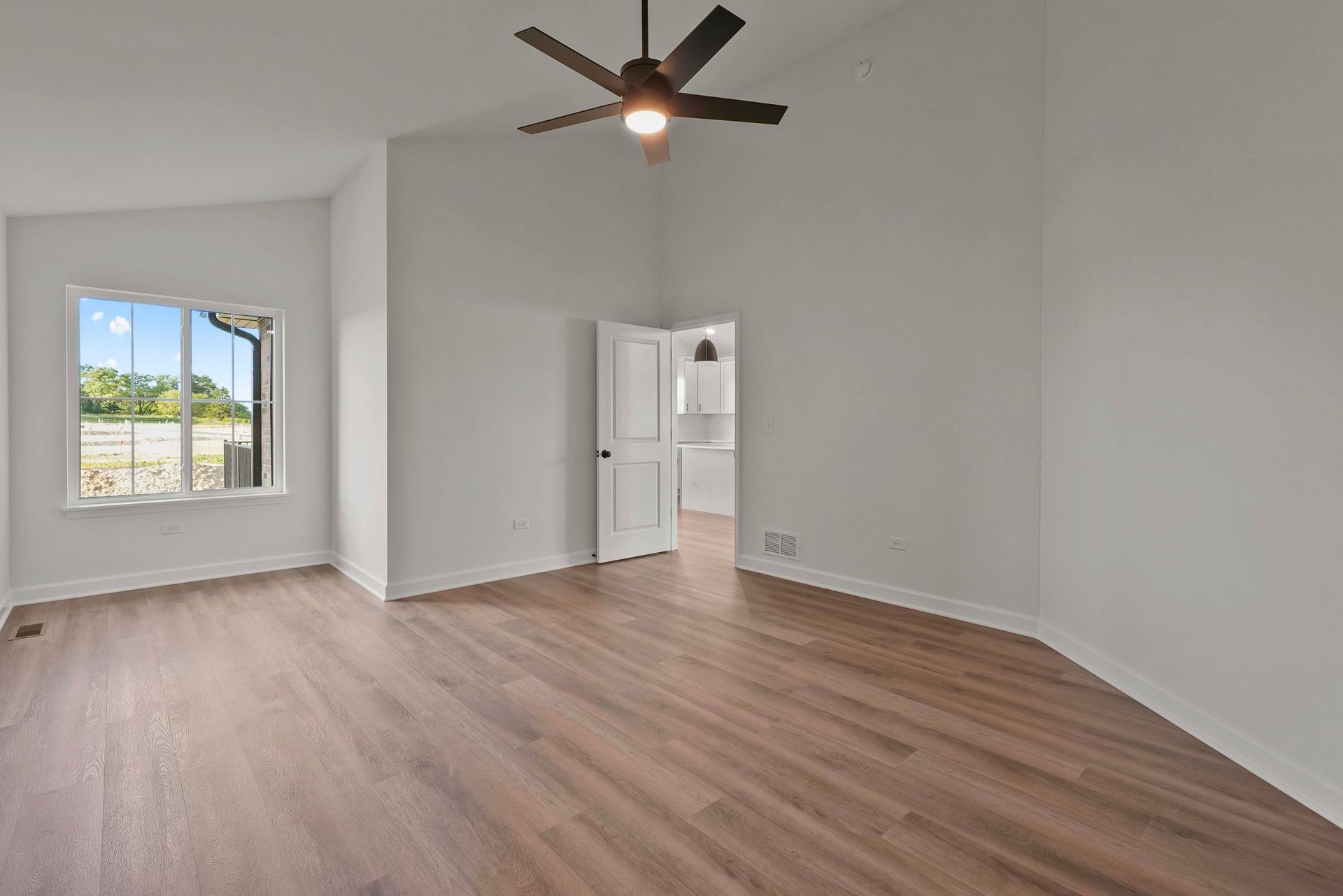 Empty modern living room with light wood floors, white walls, ceiling fan, and large window.