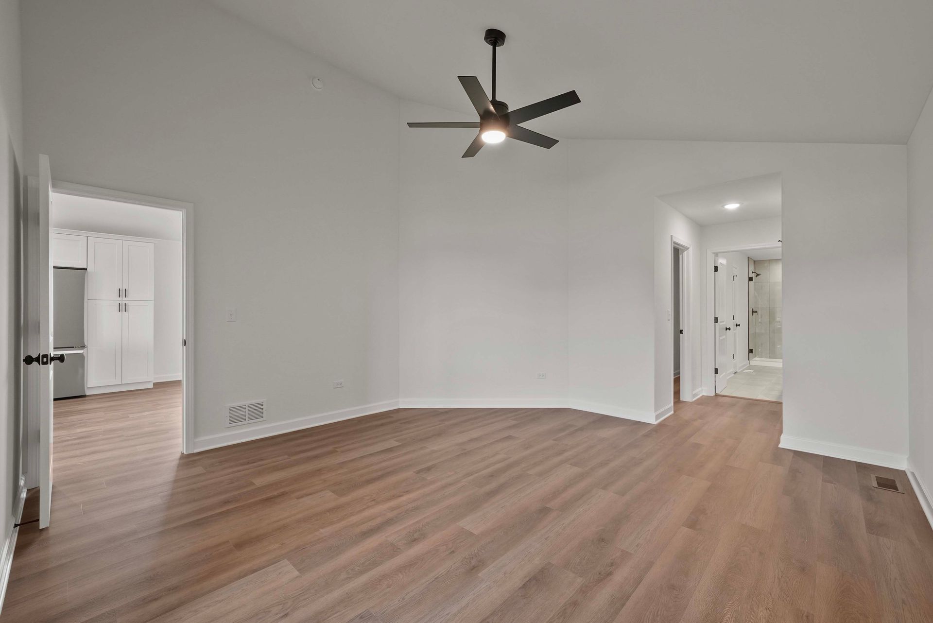 Empty modern living room with wood floors, white walls, ceiling fan, and open doorways to hall and kitchen