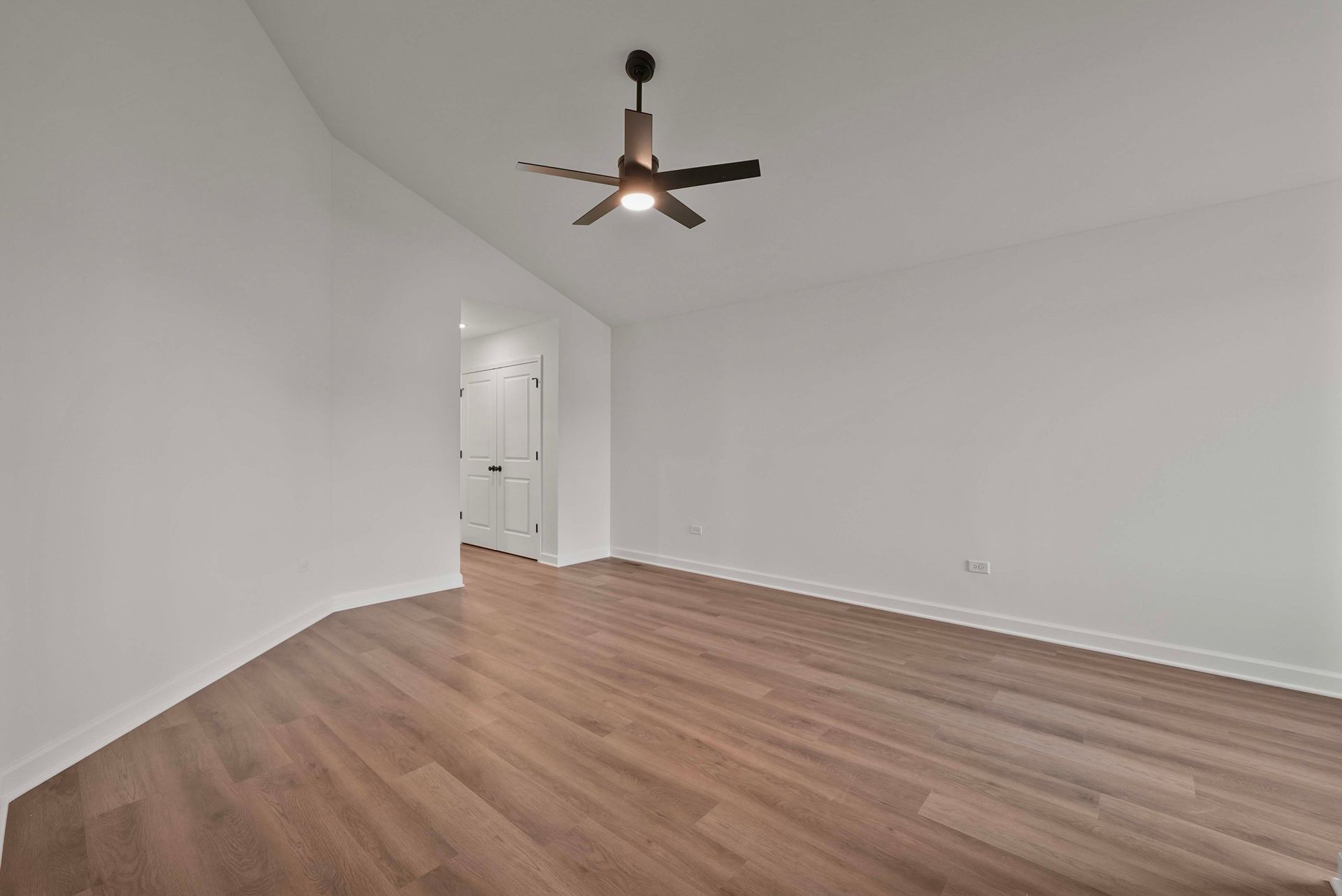 Empty white room with wood floors and a ceiling fan, viewed from a corner.
