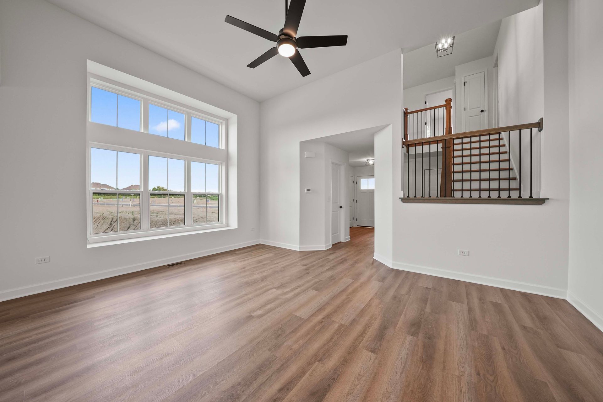 Bright empty room with large window, wood floors, ceiling fan, and stair railing opening to hallway