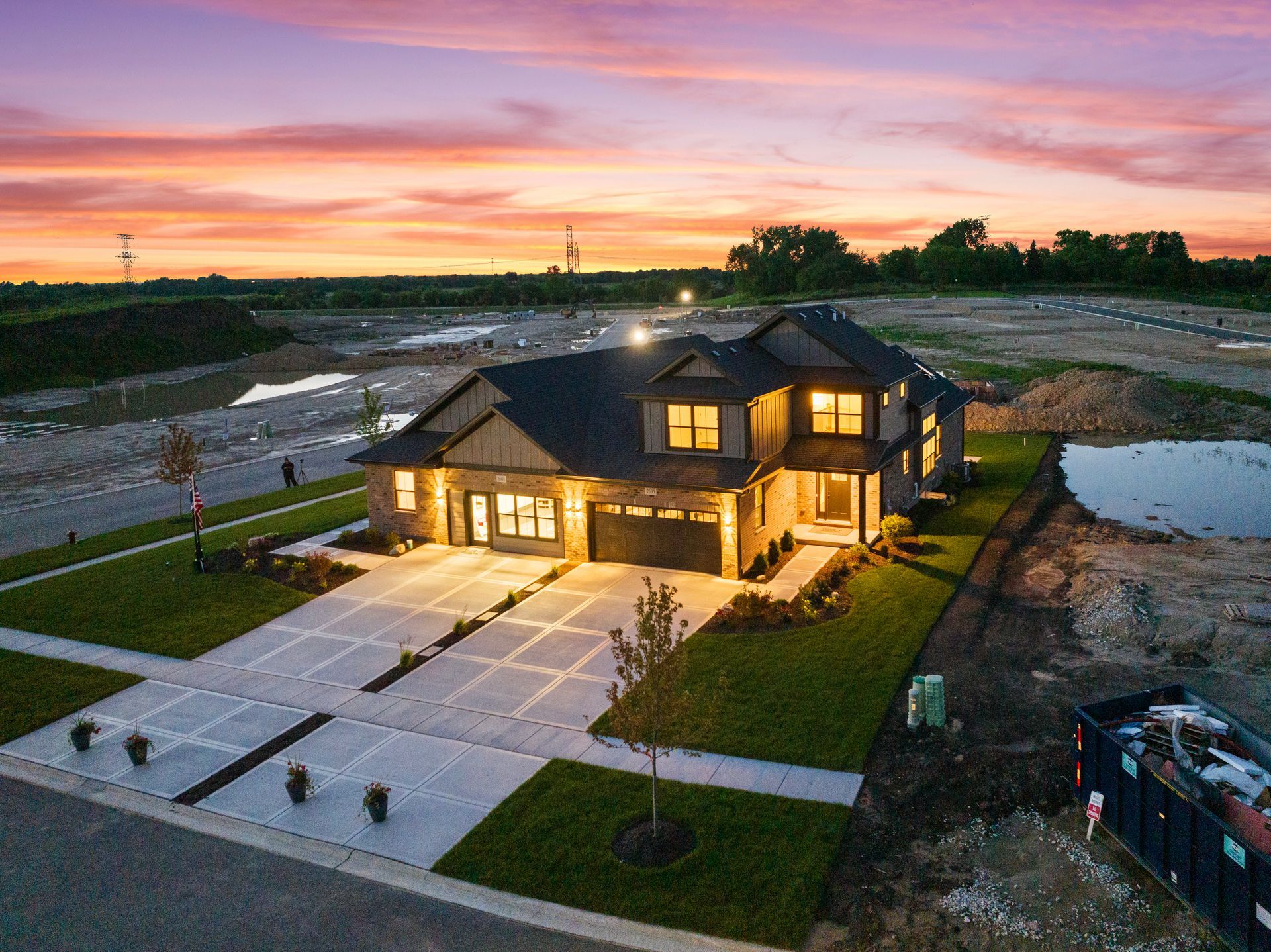 Modern two-story house at sunset, with lit windows, driveway, and a wet pond beside the property