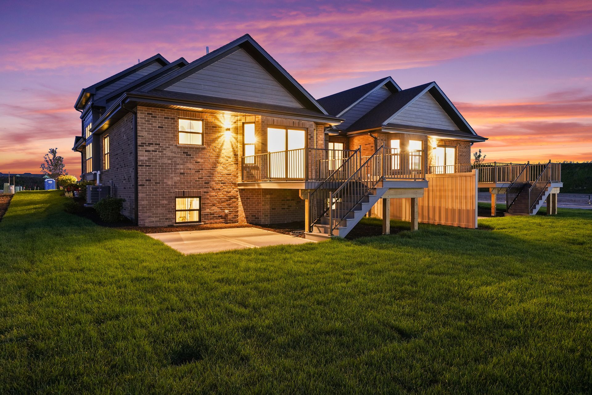 Modern stone house with warm lights at dusk, set on a grassy yard under a pink-purple sky