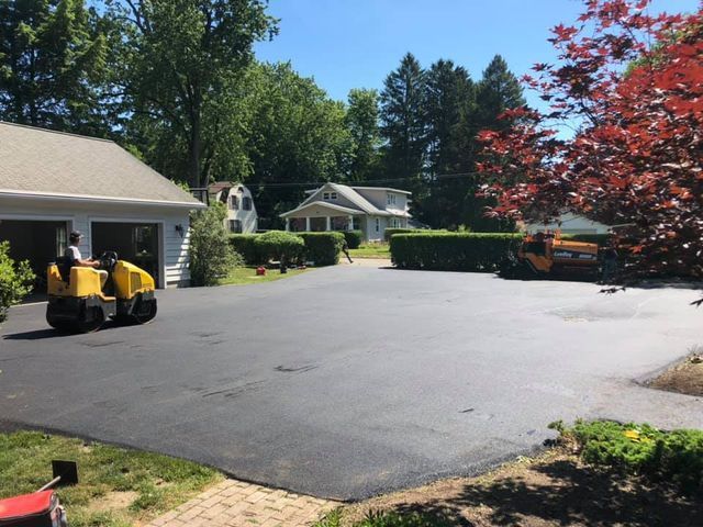 A yellow roller is rolling asphalt in a driveway in front of a house.