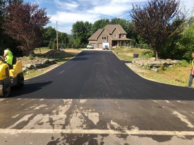 A man is driving a yellow tractor down a road in front of a house.