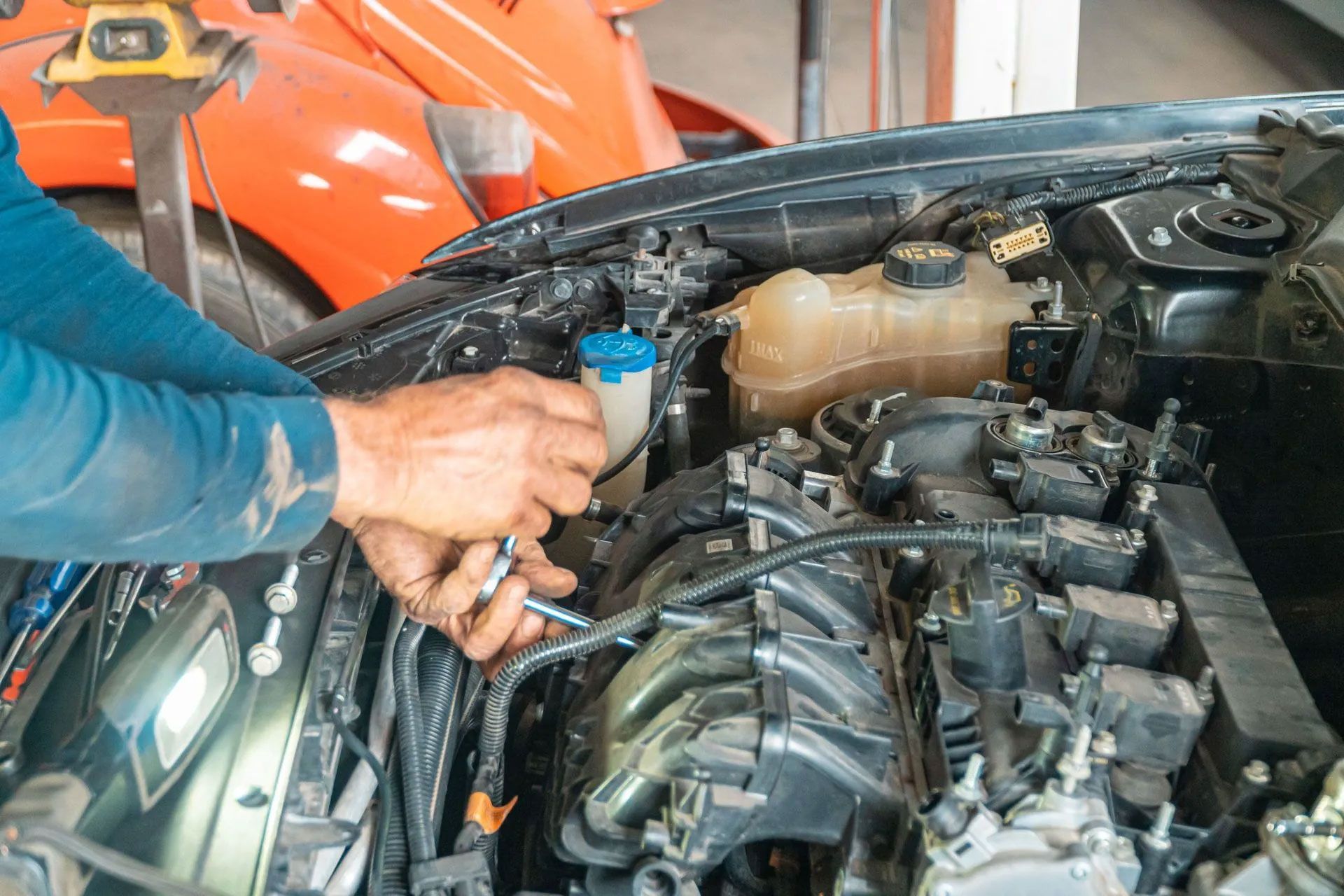 Mechanic working on a car engine with the hood open, using a tool near the battery.