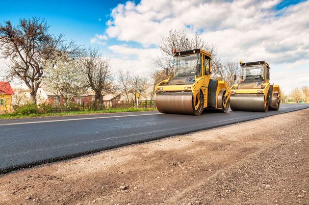Two yellow steamrollers smooth freshly laid black asphalt on a sunny road near trees and a house.