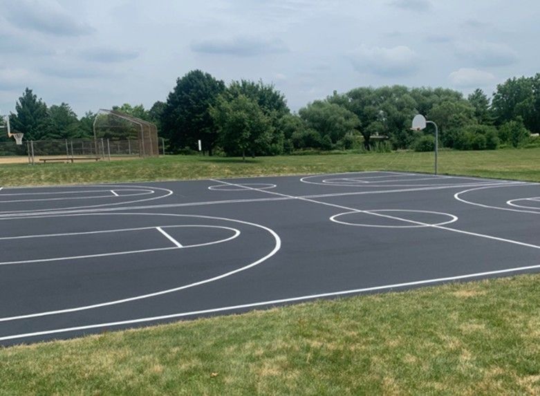 Empty outdoor basketball court with white lines, surrounded by grass and trees under a cloudy sky