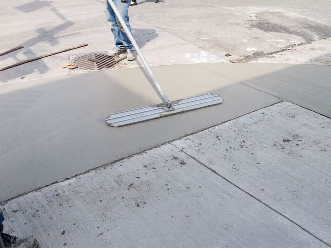 A worker uses a bull float to smooth wet concrete on a sidewalk construction site.
