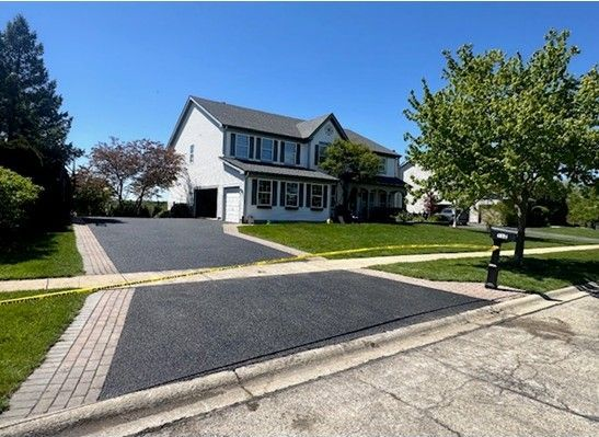 Suburban house with a wide dark driveway, green lawn, trees, and a blue sky.