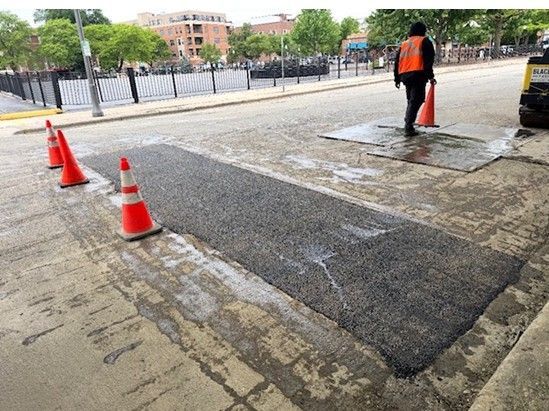 Road repair crew patching a sidewalk, with orange cones marking the work area