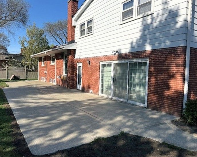 Concrete patio beside a brick-and-white house with a sliding glass door and covered porch on a sunny day