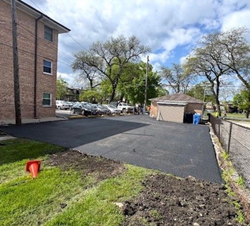 Freshly paved black asphalt beside a brick building, with grass, trees, and a small shed in the background