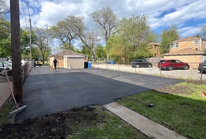 Apartment parking lot with two people near a shed, cars along the curb, and trees under a partly cloudy sky
