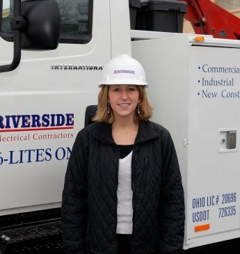 Woman in hard hat stands by Riverside Electrical truck, smiling.