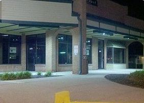 Exterior of a building with large glass windows, a covered walkway, and a paved pathway at night.