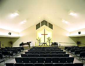 Church interior with rows of chairs facing a stage with a cross, piano, and altar.