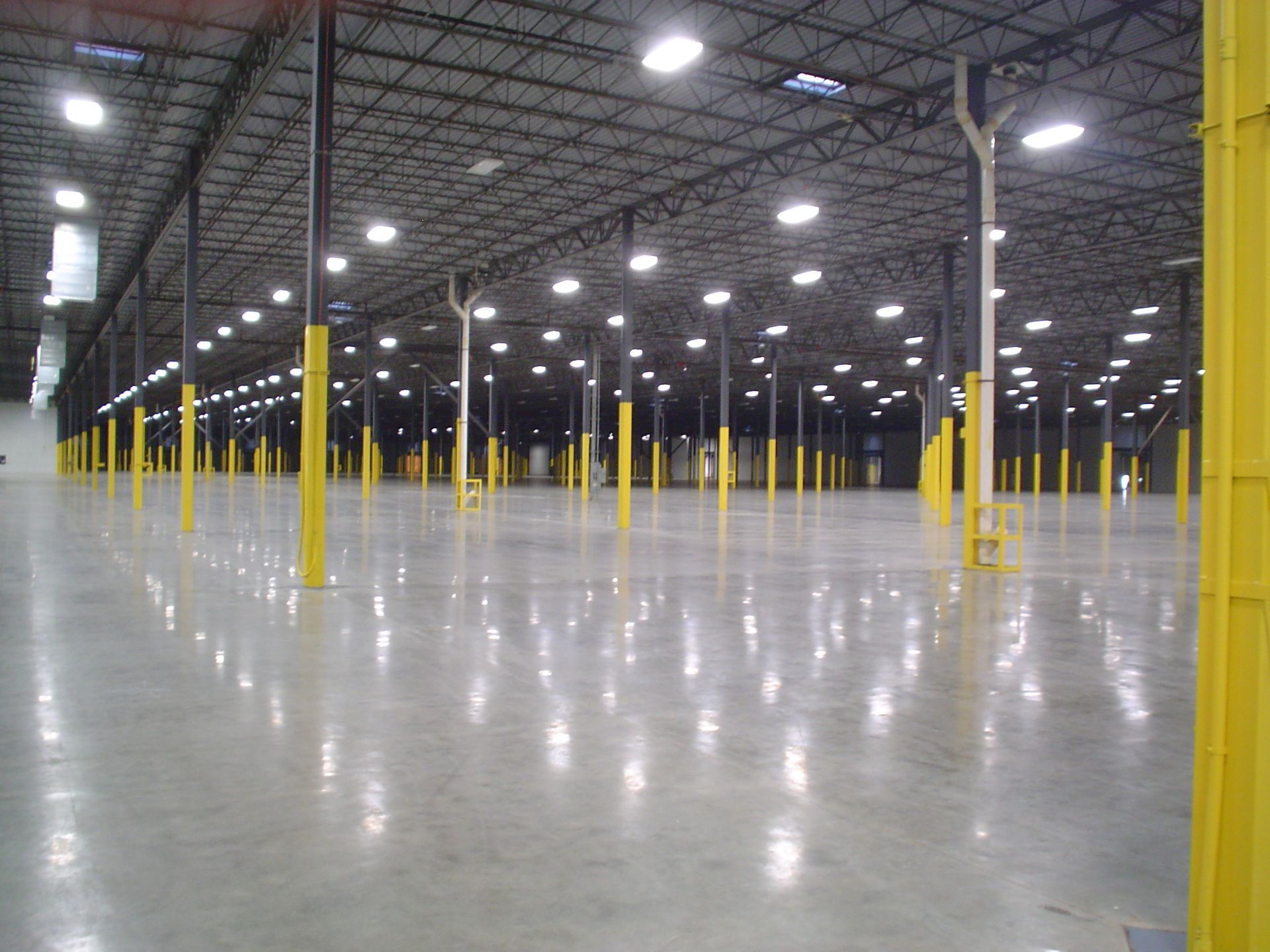 Empty warehouse interior with reflective concrete floor, yellow columns, and overhead lights.