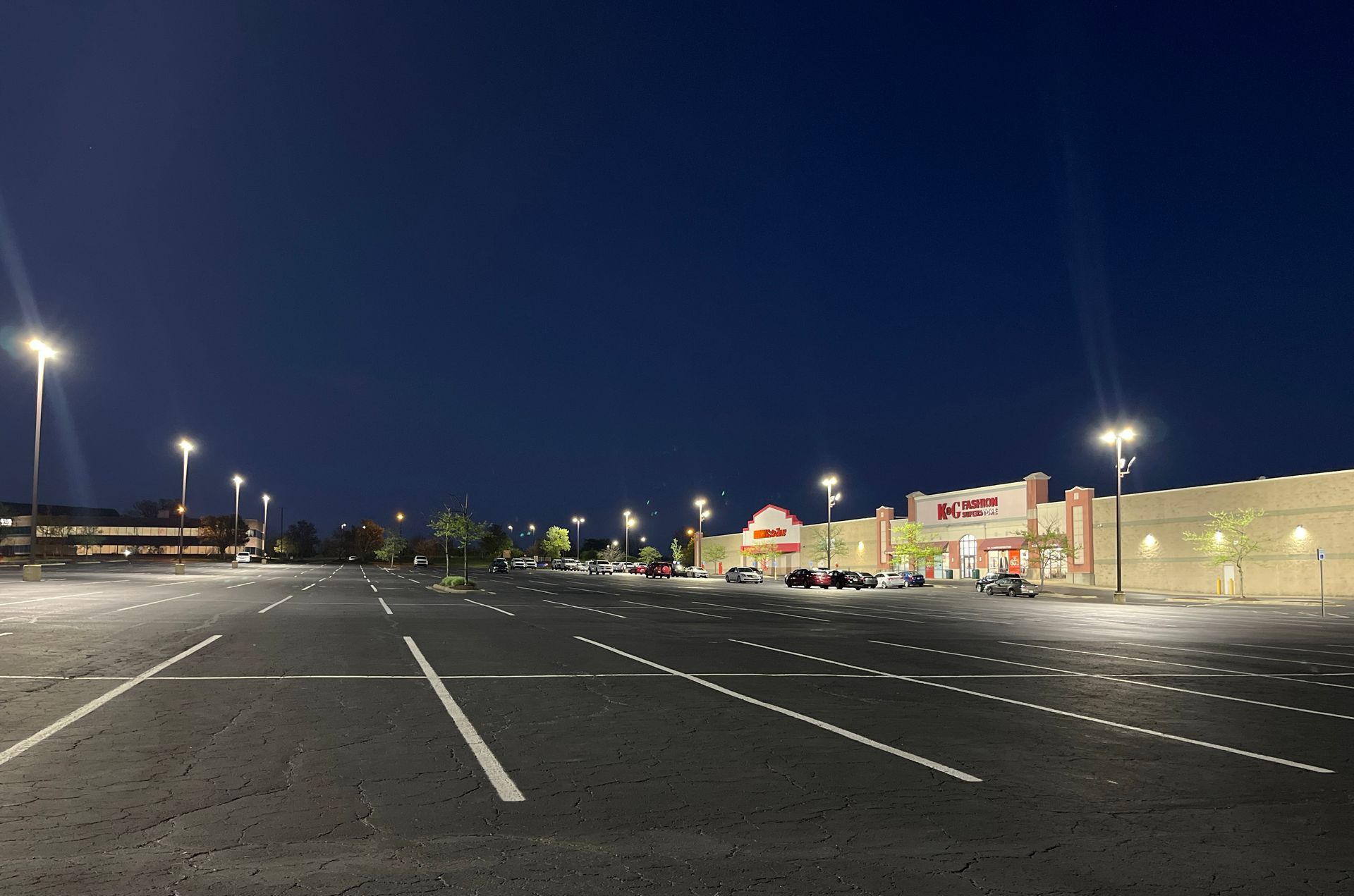 An empty asphalt parking lot at dusk with streetlights illuminating a building with storefronts.