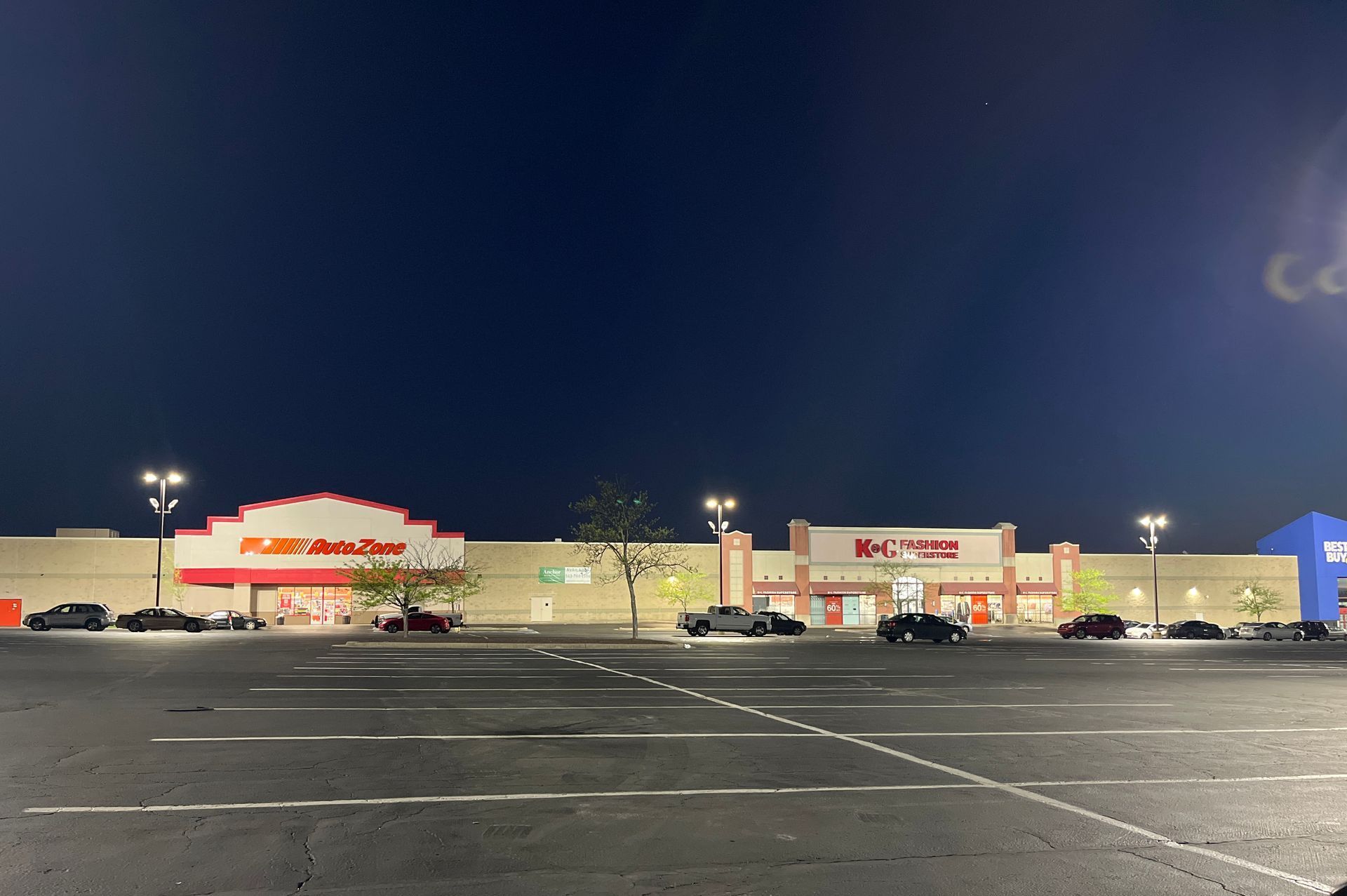 A nighttime photo of a nearly empty parking lot in front of a strip mall with several businesses, including an auto parts store.