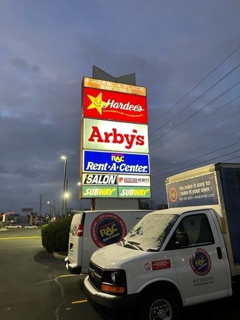 A lit signpost for a shopping center at dusk, with Hardee's, Arby's, Rent-A-Center, Subway, and salon. Trucks are parked in front.