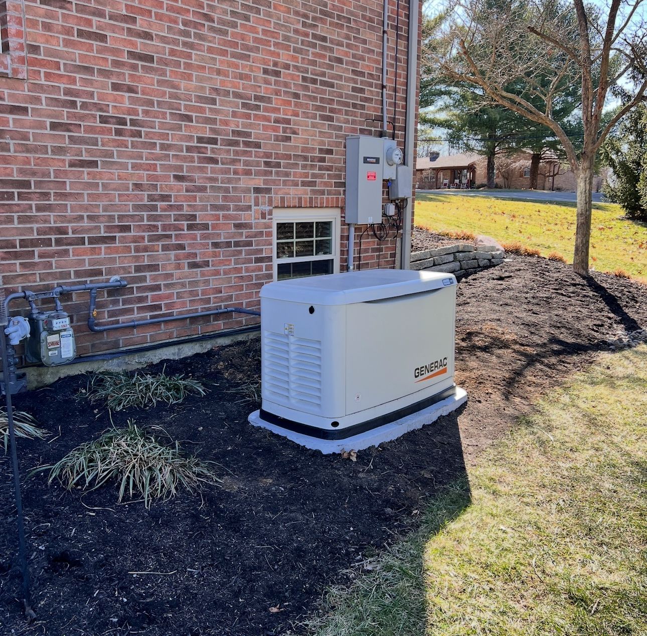 A standby generator sits outside a brick home, near a utility box and landscaped bed.