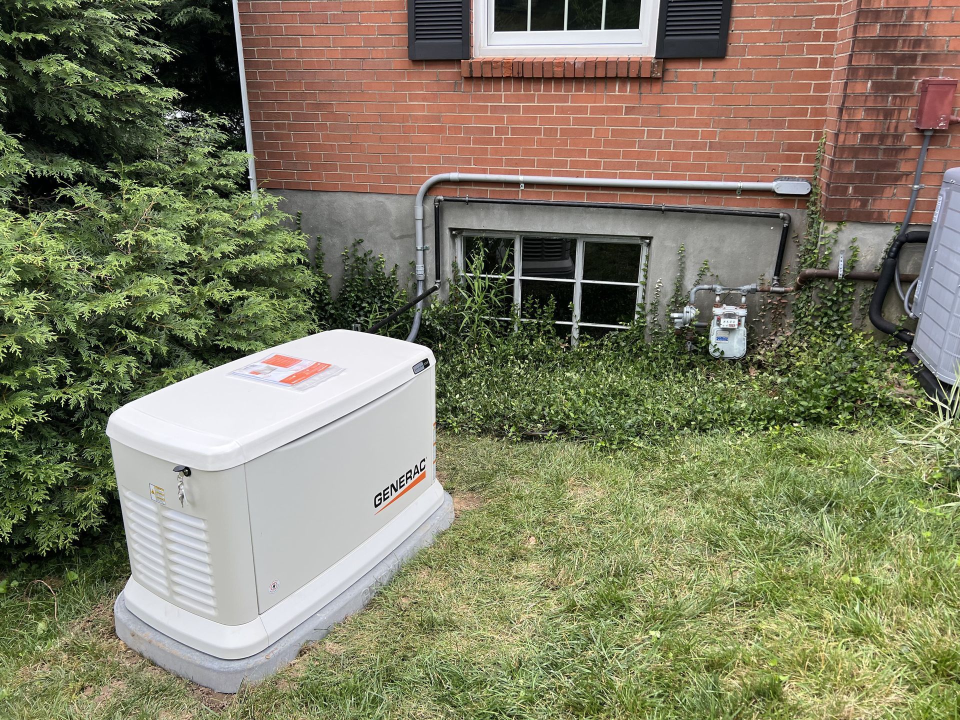 Generator outside a brick building, near a window and overgrown foliage.