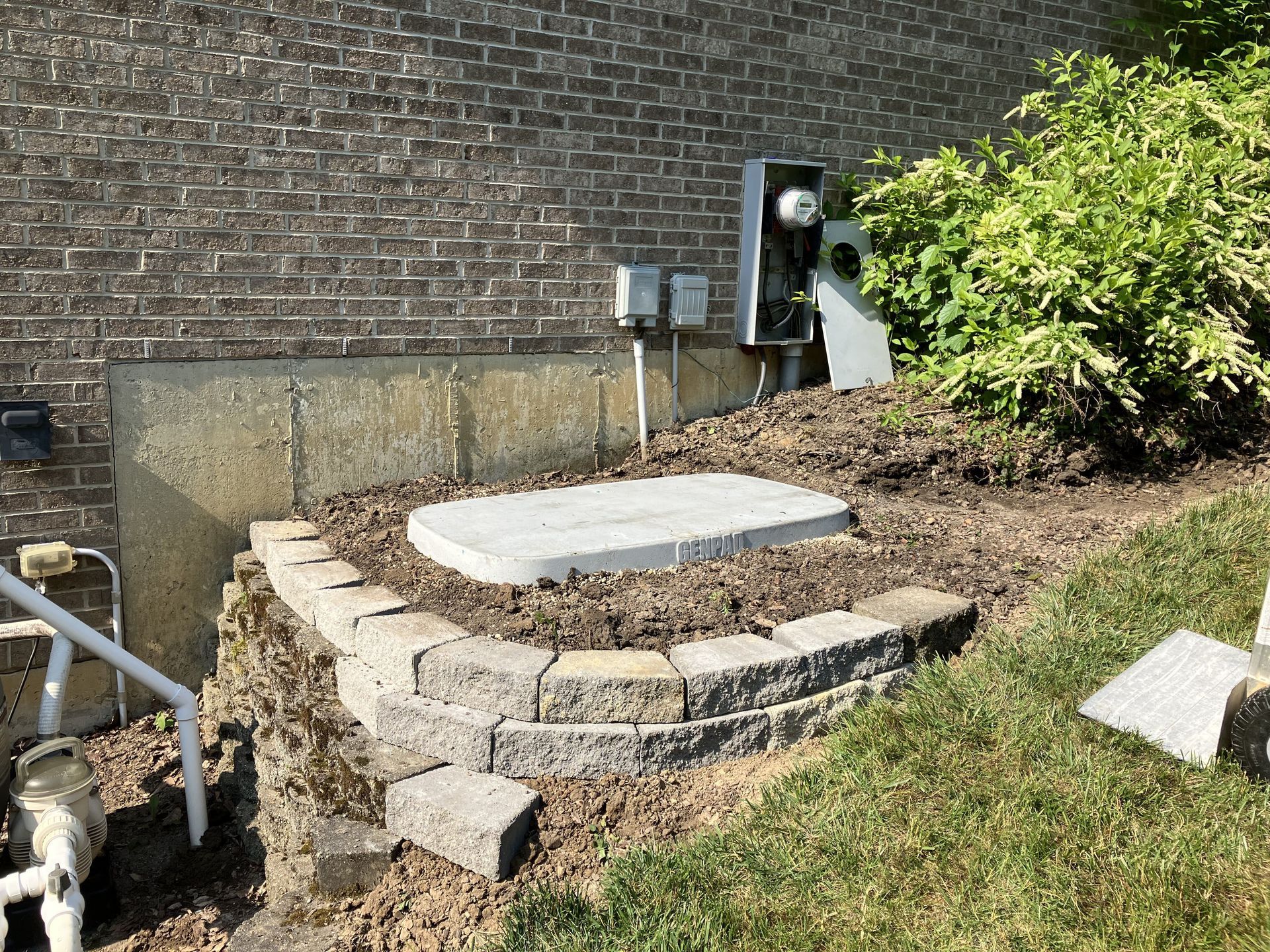 Brick retaining wall surrounding a concrete lid and electrical boxes, near a building with brick and bushes.