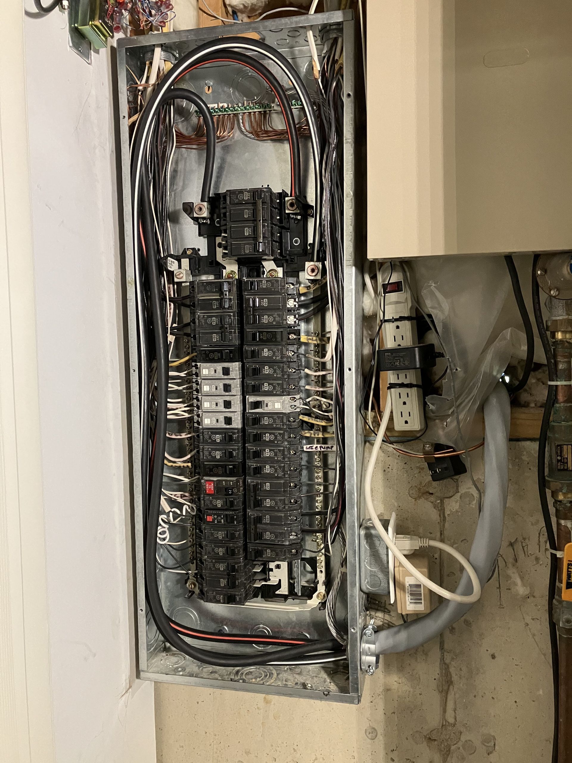 Open electrical panel with many circuit breakers and wires against a wall.