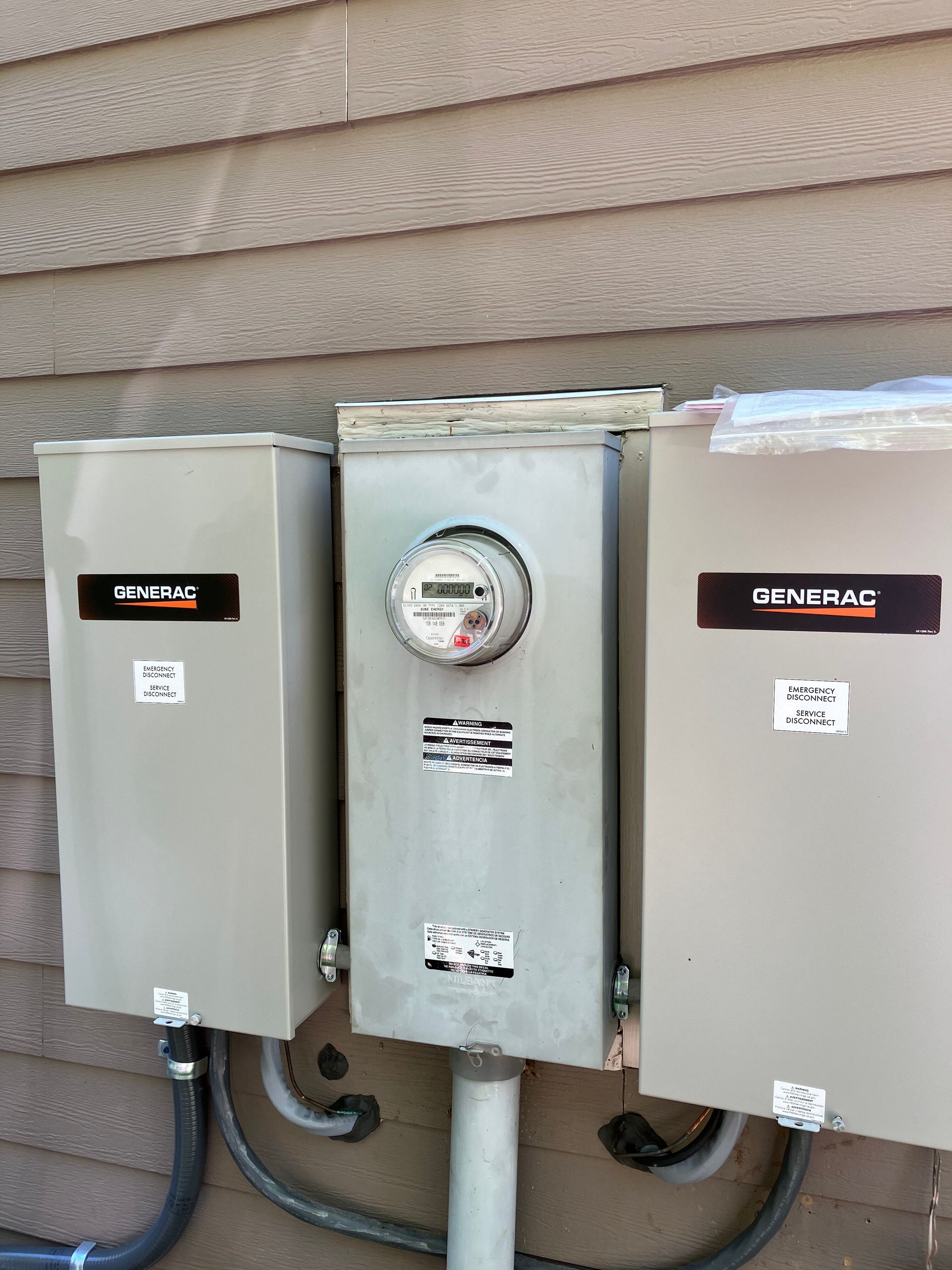 Electrical meters and boxes on a brown siding wall. The boxes are gray and bear the Generac logo.