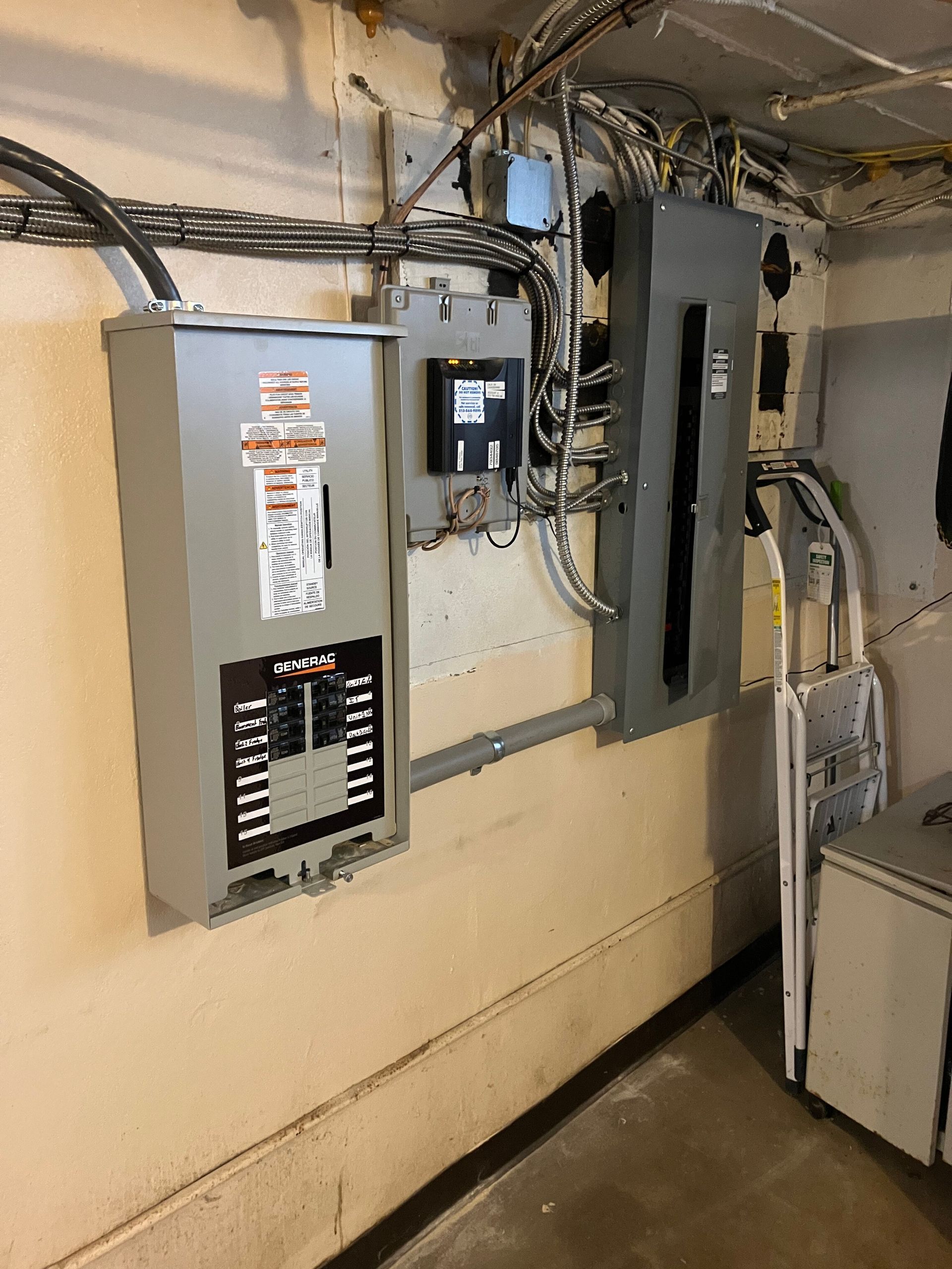 Electrical panel in a basement. Gray metal boxes mounted on a beige wall with conduits and wires.