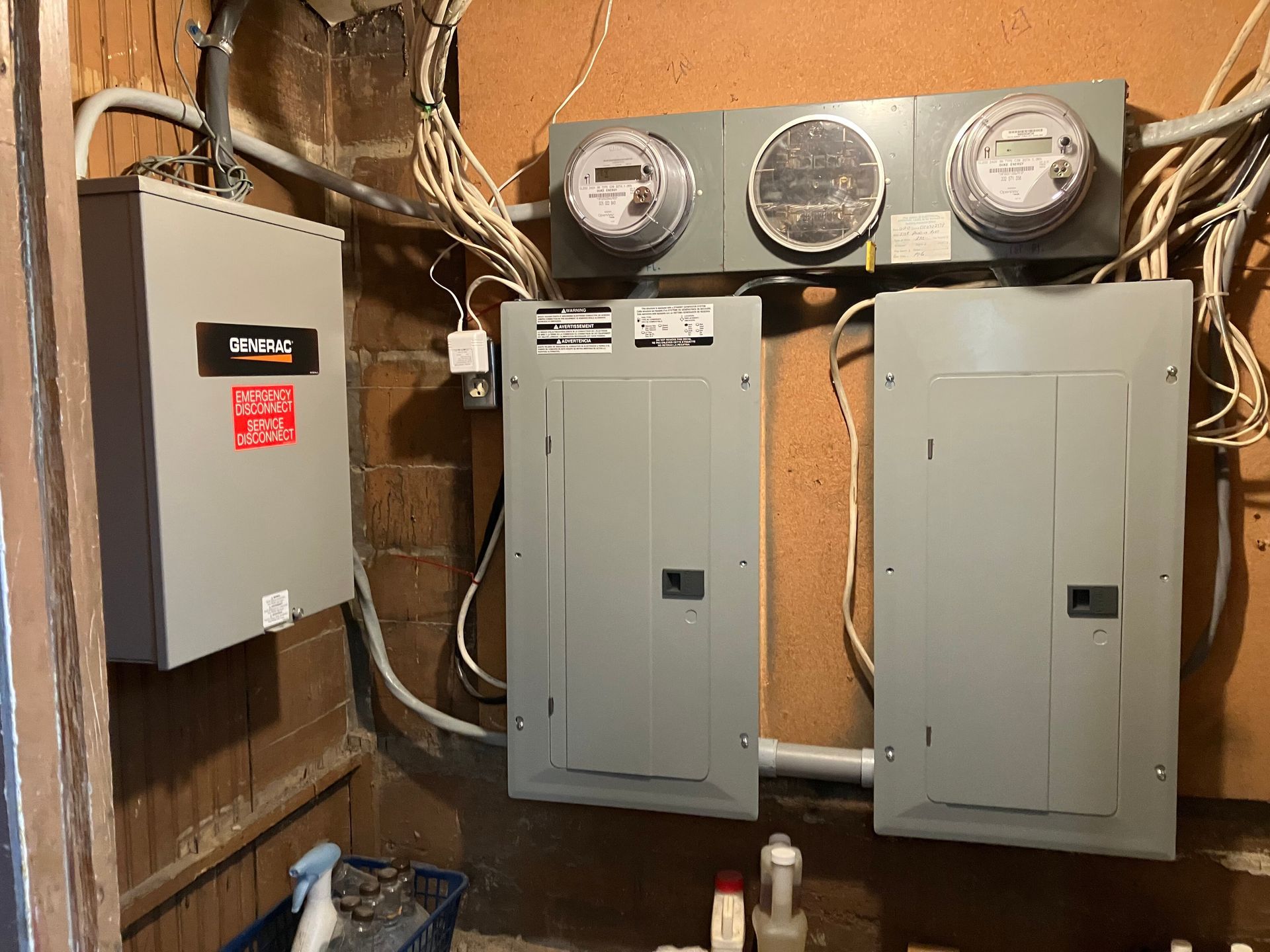 Electrical panel setup: three meters above two gray breaker boxes, connected by conduits and wires on a wood paneled wall.
