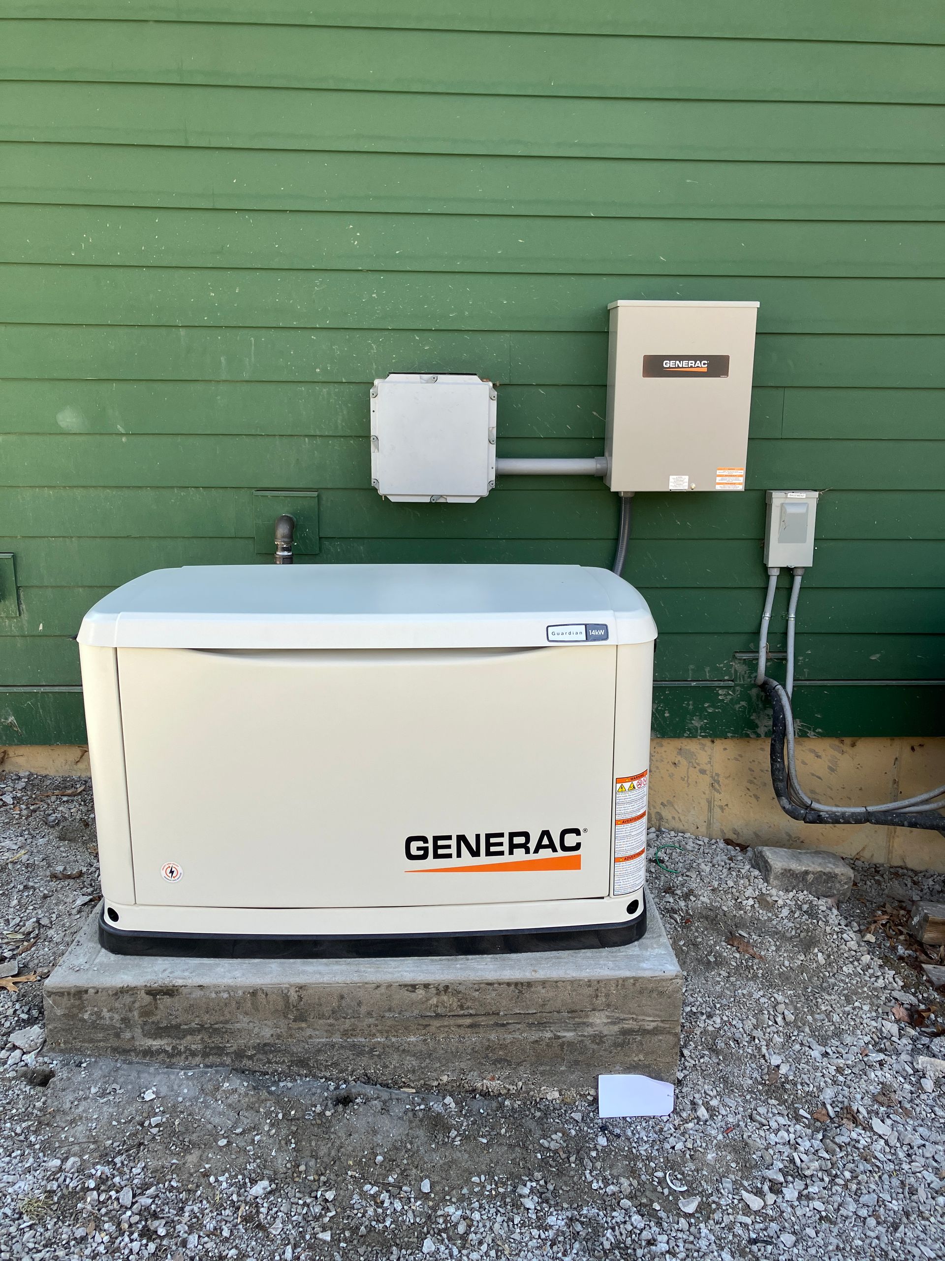 A Generac generator outside a green building. Includes control boxes and concrete pad on a gravel base.
