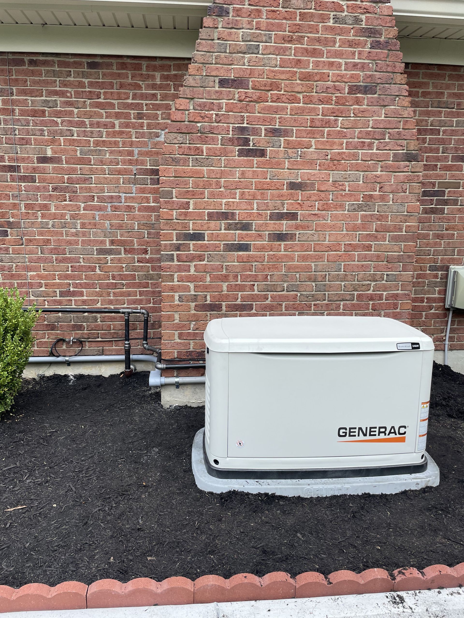 A white Generac generator on a concrete pad next to a brick wall and mulch.