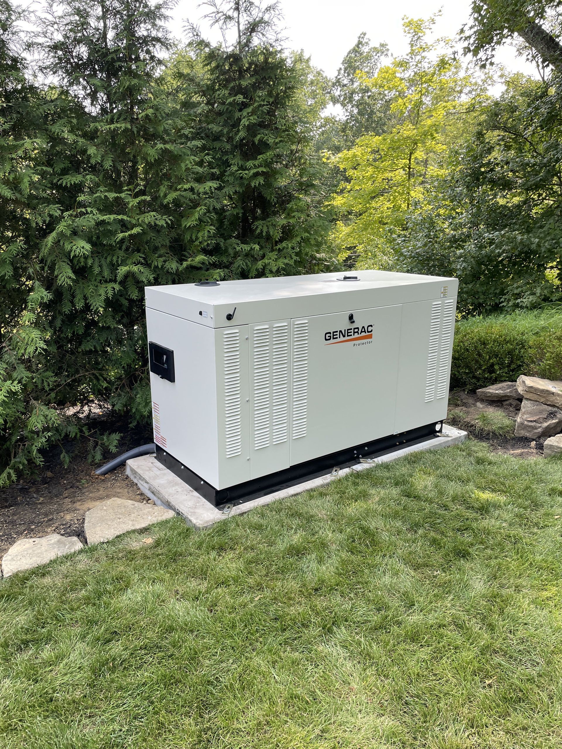 A white generator on a concrete pad in a grassy yard, surrounded by trees and bushes.