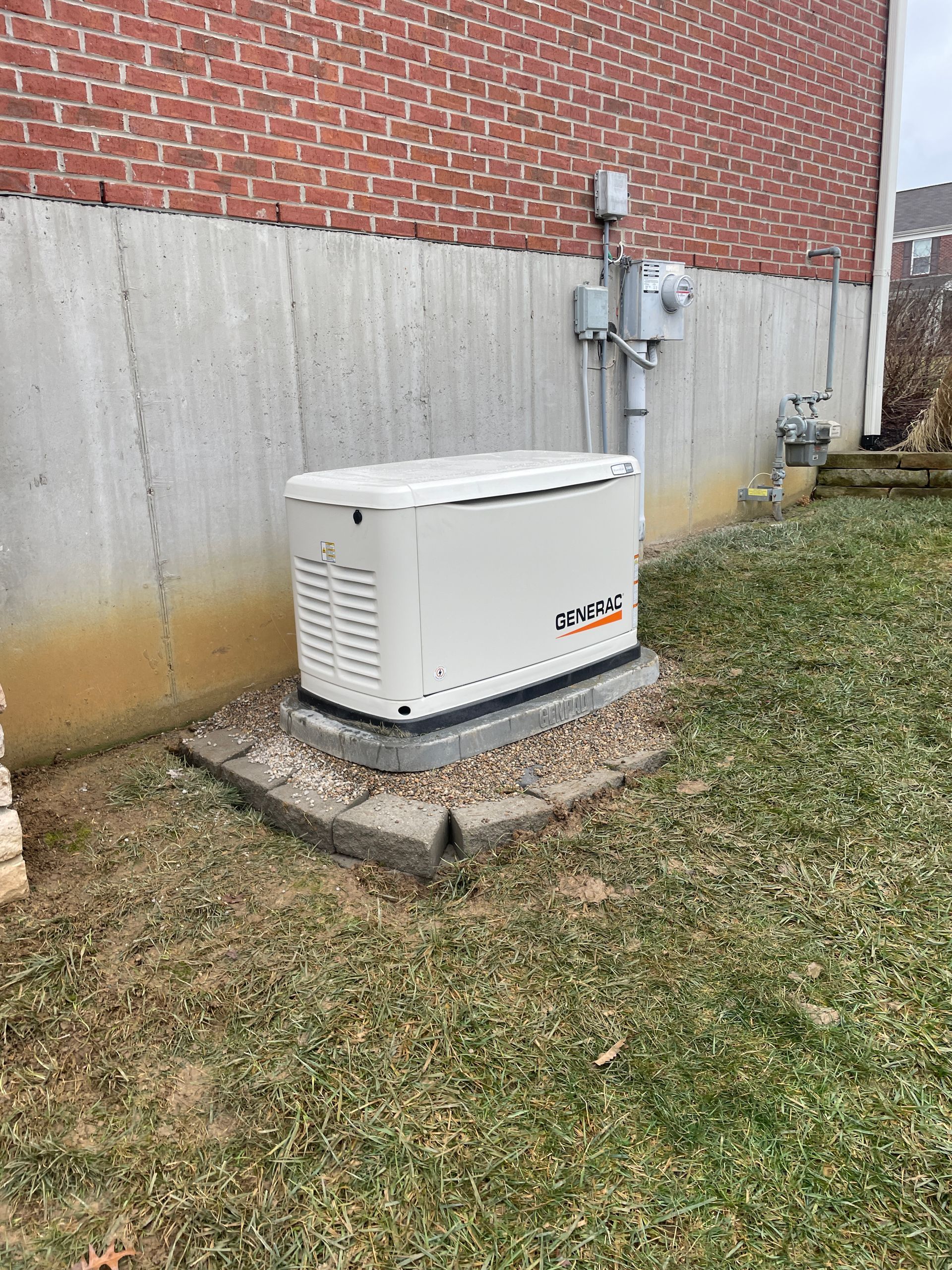 White generator on a concrete base next to a brick wall and utility pipes. Grass and gravel surround.