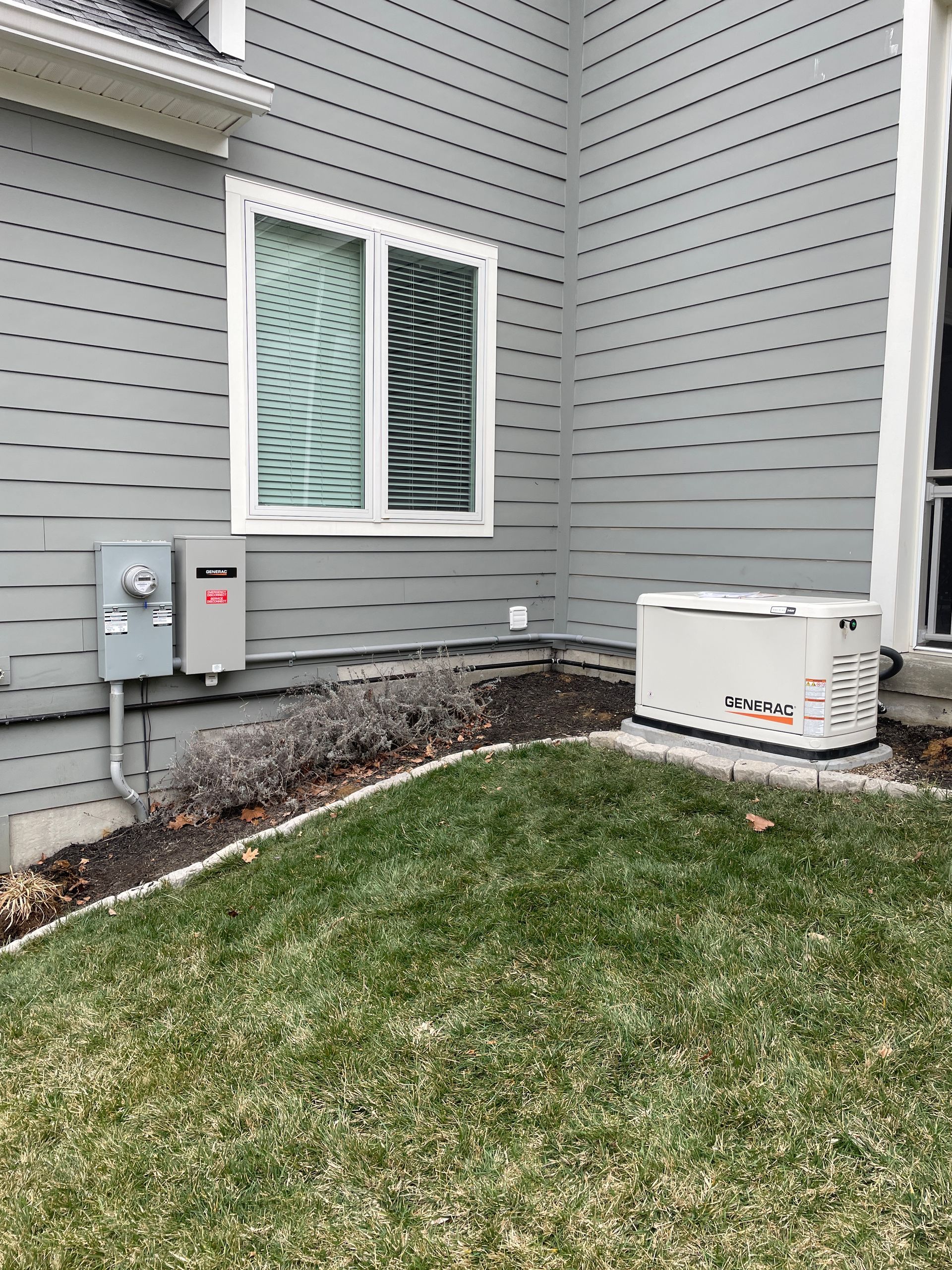 A house exterior with gray siding, a window, and a generator unit next to electrical boxes in the grass.