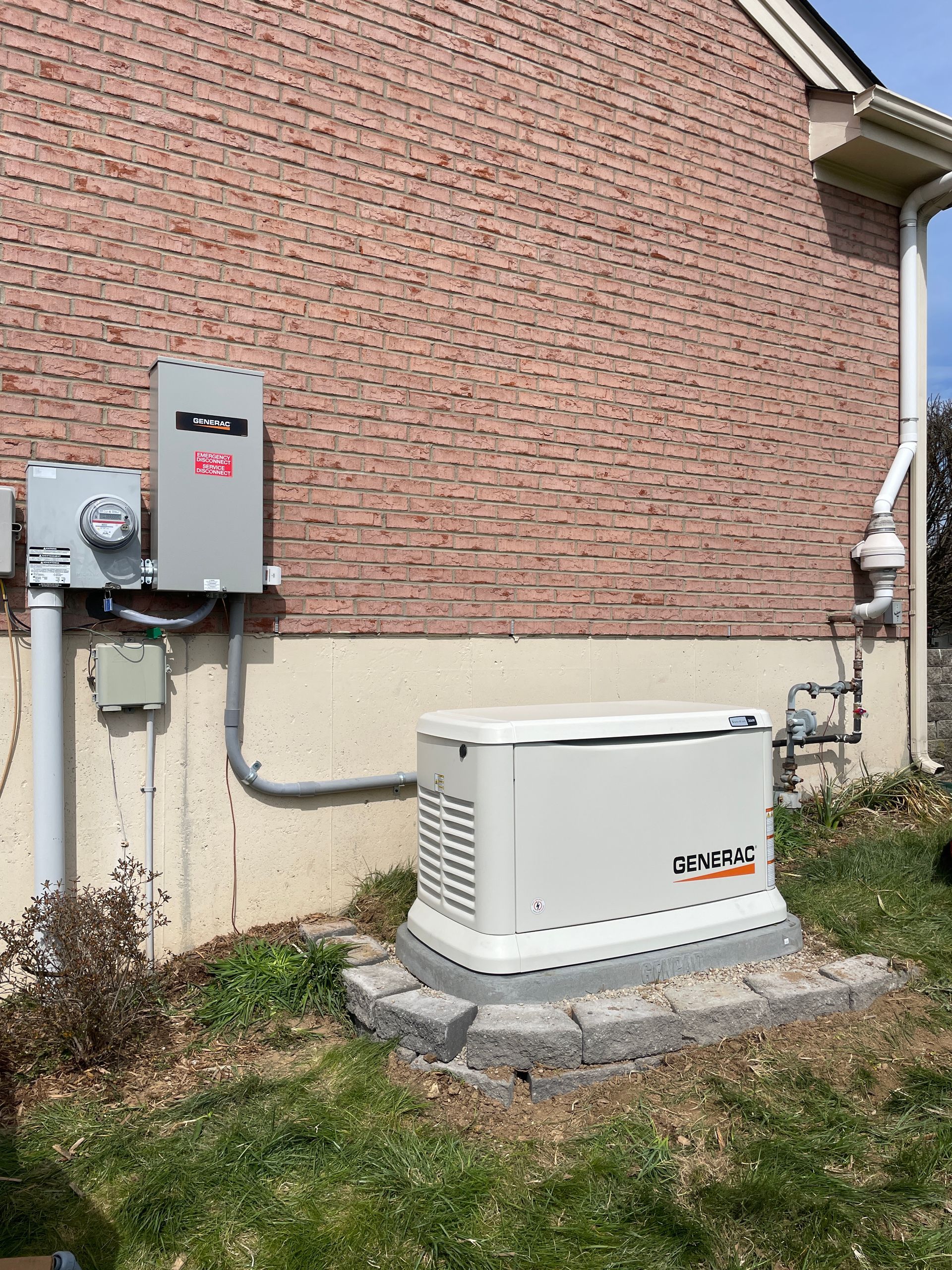 A standby generator installed against a brick building. A gray electrical box and meter are mounted above it.