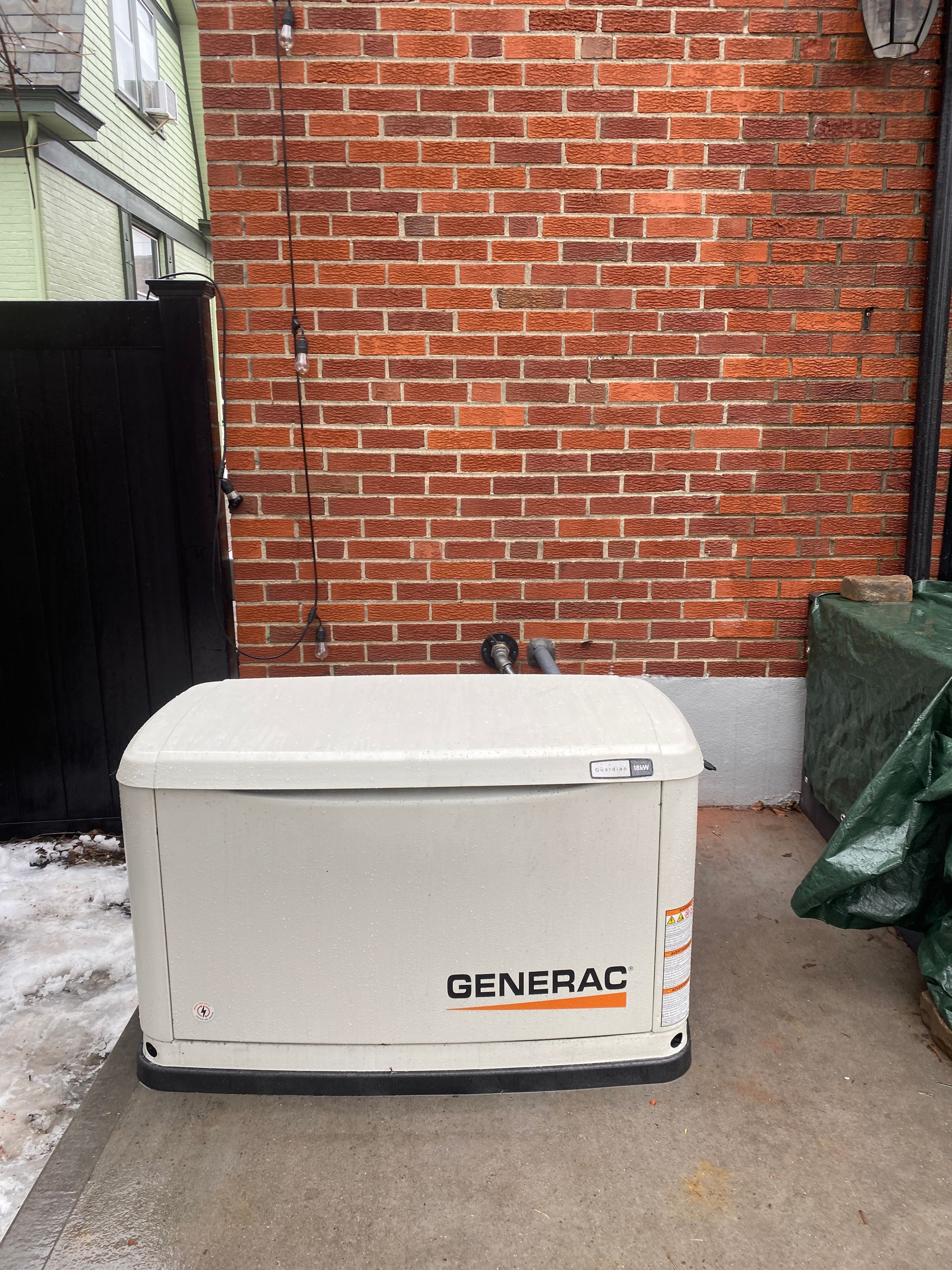 Generac generator against a brick wall; a black gate and snow on the ground are also visible.
