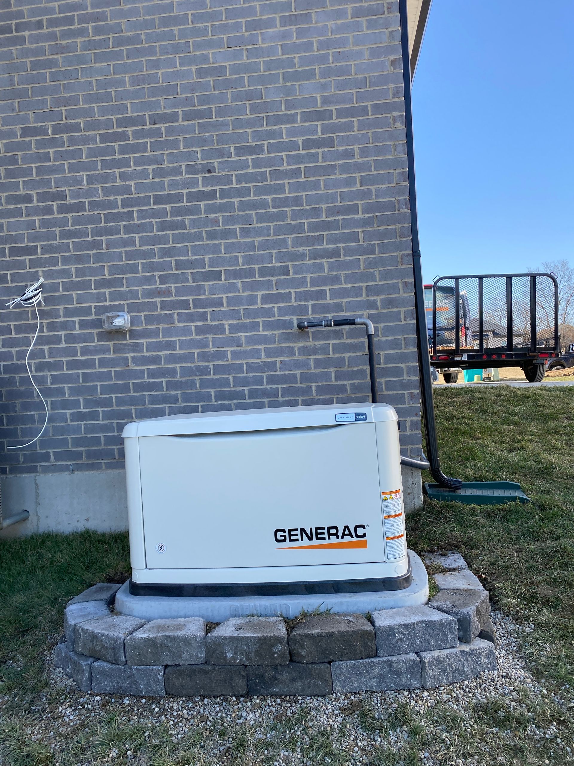 Generac generator on a stone base, next to a brick building, under a clear sky.